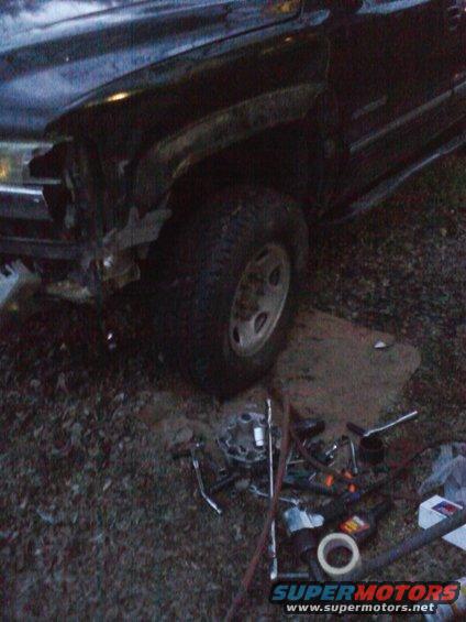 Dark SUV with damaged front fender and bumper, parked on dirt with tools scattered around the front wheel