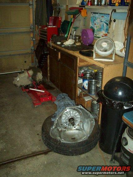 transmission on tire, another transmission on red jack, workbench with tools and fan in garage