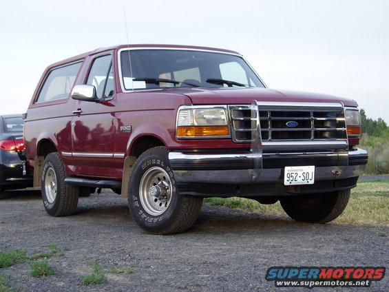 Maroon Ford Bronco parked on gravel, front 3/4 view. Another car is partially visible to the left