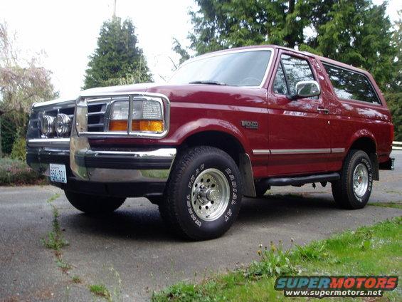 Red 1993 Ford Bronco parked next to a white truck. The Bronco has chrome wheels and a side step