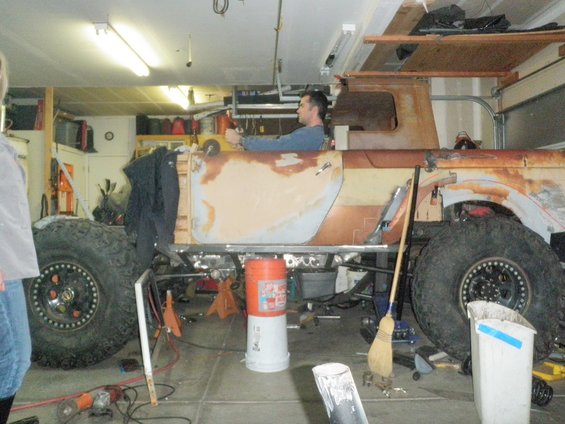 Side view of a rusted pickup truck body with large off-road tires, undergoing restoration