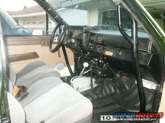 Interior view of a truck with bucket seats, roll cage, and dashboard. A blue vehicle is visible through the driver's side window