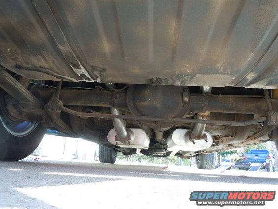 Undercarriage of a car showing the rear differential, exhaust pipes, and fuel tank. A blue truck is visible in the background