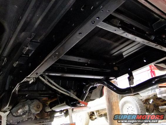 Underside of a vehicle frame with exhaust pipe and drivetrain components visible. Another vehicle is partially seen in the background