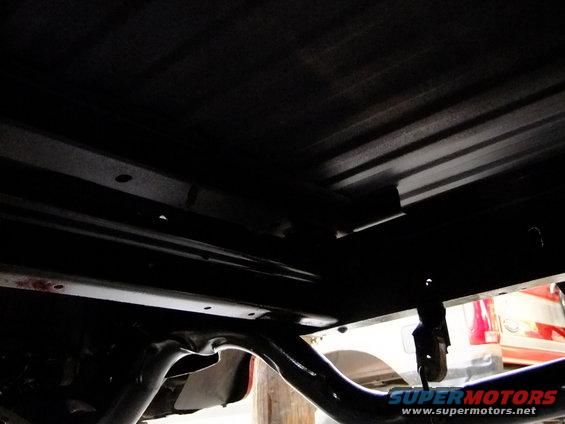 Underside view of a vehicle's frame and suspension components. A red taillight from another vehicle is visible in the background