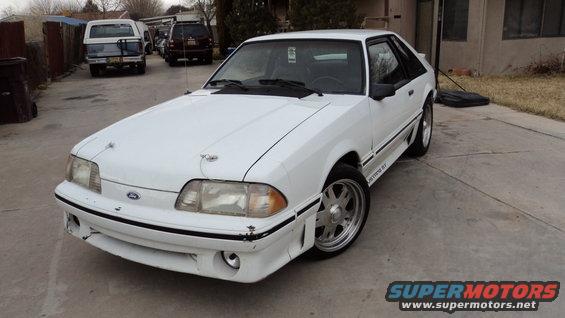 White 1987 Ford Mustang GT with aftermarket wheels, hood pins, and body kit parked on a concrete driveway