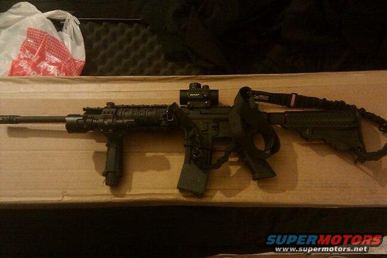 AR-15 style rifle with red dot optic and black sling, resting on a cardboard surface