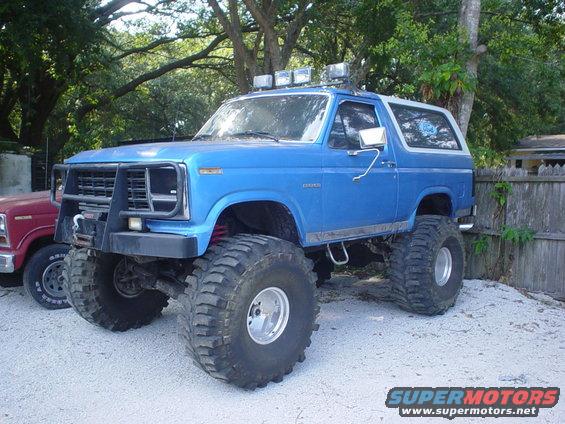 Blue lifted Ford Bronco with off-road tires and roof lights, parked next to a red truck