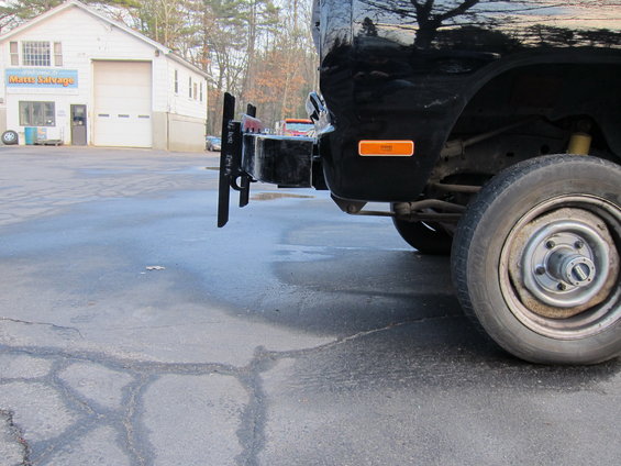 Black pickup truck's rear bumper with a tow hitch and orange reflector. Building in background