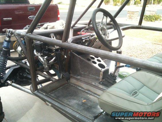 Custom buggy interior with roll cage, bucket seats, steering wheel, and shifter. A red Ford pickup is visible in the background