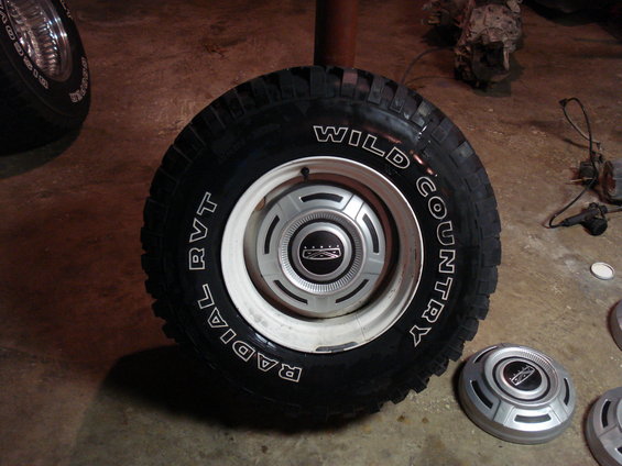 Close-up of a Wild Country radial tire on a white rim with a Ford hubcap. Another hubcap is on the floor