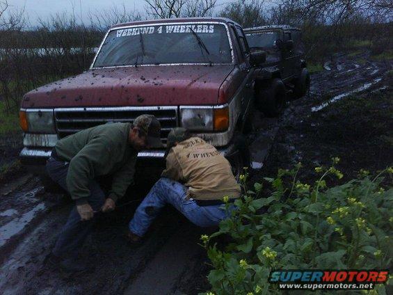 1989 Ford Bronco stuck in mud with two men winching it, another vehicle behind