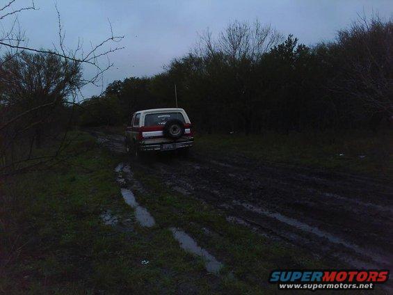 1989 Ford Bronco driving on a muddy dirt road with trees in the background