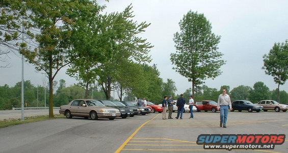 Parking lot scene with a line of cars and a few people standing near them