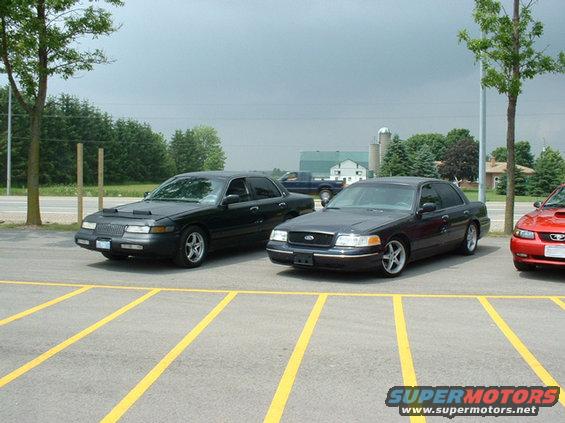 Two dark Ford Crown Victorias parked in a lot, one with a hood cover. A red car is partially visible on the right