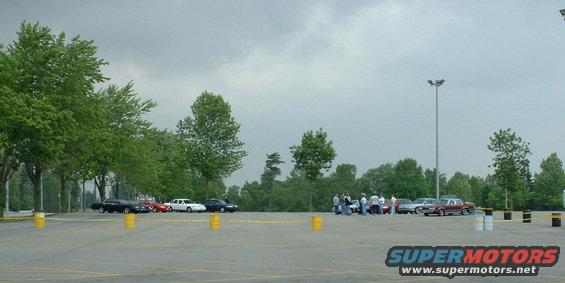 Cars parked in a lot with a group of people standing near some of them under a cloudy sky