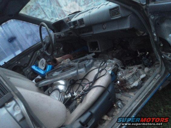 Car interior with damaged dashboard, torn seats, and scattered engine parts. Blue intake pipe visible