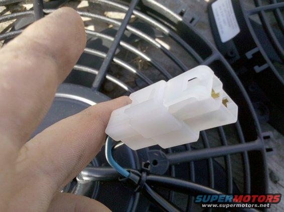 Close-up of a white electrical connector with blue wire attached, held by a finger, in front of a black fan grille