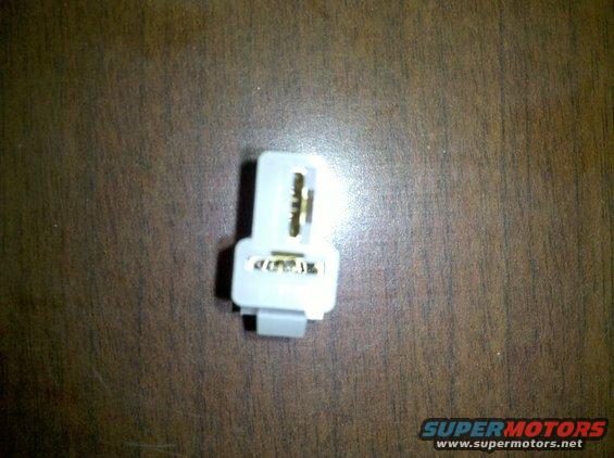 White electrical connector with gold terminals on a wood-grain surface. Supermotors.net logo in the bottom right