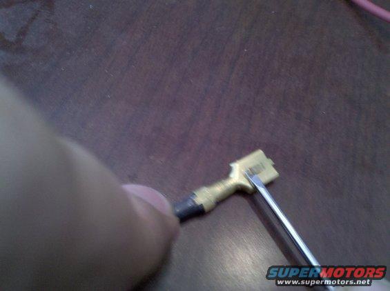 Close-up of a hand holding a small electrical connector with a screwdriver tip touching it. Supermotors.net logo visible