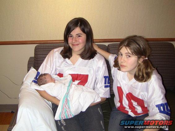 Two girls and a baby wearing New York Giants jerseys sit on a couch