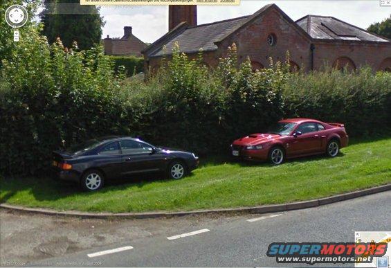 red Ford Mustang Cobra and black Toyota Celica parked on grass next to a brick building