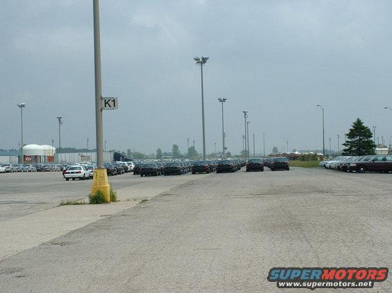 Rows of dark sedans and a few white cars parked in an outdoor lot under a cloudy sky. A sign reads 'K1'