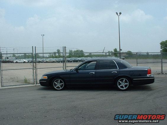 Dark blue 2000 Ford Crown Victoria parked on asphalt, side view. Multiple cars in background behind fence