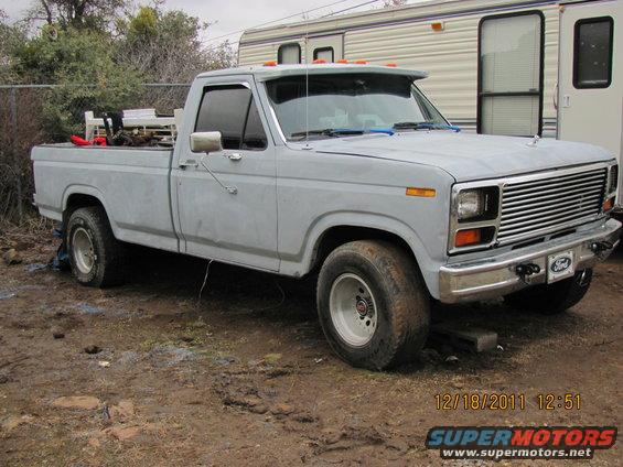 Gray Ford F-150 pickup truck with aftermarket visor and chrome grille, parked on dirt. RV trailer behind it