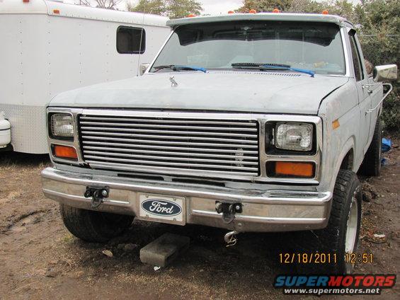 Gray Ford F-150 truck front view, chrome grille, Ford badge, some damage, parked outdoors