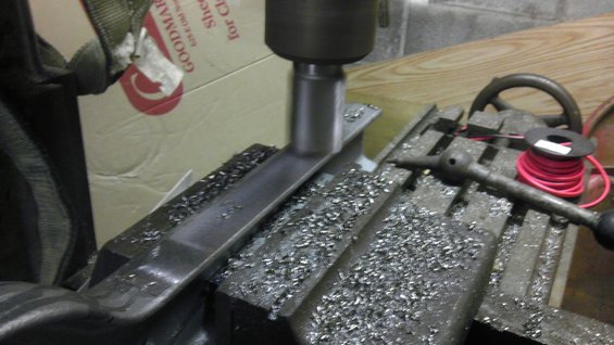 Milling machine cutting a metal part, with metal shavings on the table. A box with 'GOODMARK' is in the background
