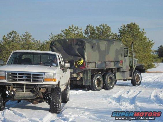 White Ford F-Series pickup truck towing a camouflaged military cargo truck in the snow