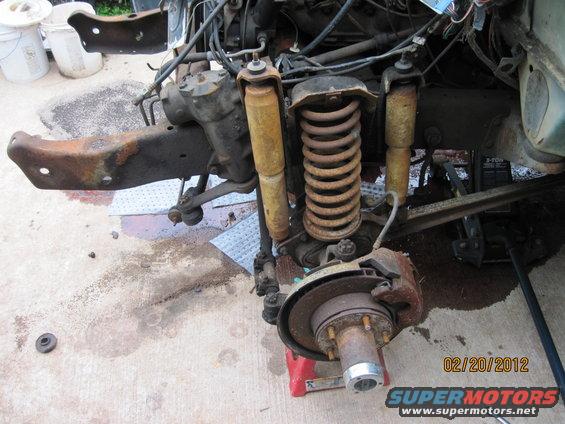 Front suspension and steering parts of a vehicle, likely undergoing repair. Shows coil springs, shocks, brake rotor, and steering gear