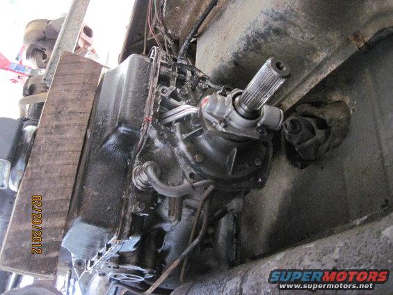 Underside view of a vehicle's transmission and driveshaft with wooden support and visible date stamp