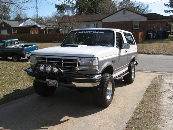 1994 Ford Bronco, lifted with aftermarket bumper, lights, and hood scoop. Blue pickup truck in background
