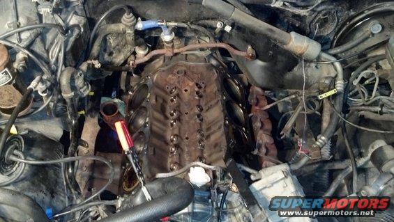 Engine bay with a V8 engine block, exhaust manifolds, and various hoses and wires. A red and silver tool is visible near the engine