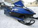 1987 Polaris Indy 600 - A blue Yamaha SRX snowmobile sits in the snow, with a covered vehicle and houses in the background.