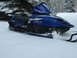 1987 Polaris Indy 600 - A blue Yamaha SRX snowmobile sits in a snowy landscape, with trees visible behind it.