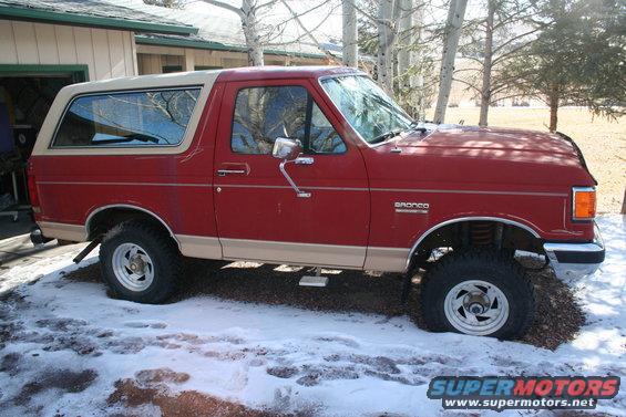 Red Ford Bronco with tan removable top, lifted suspension, parked on gravel and snow