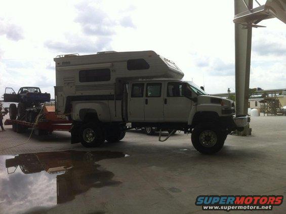 White custom camper truck with four doors, towing a trailer with a blue pickup truck on it