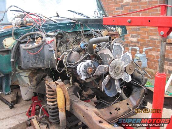 Green vehicle engine and chassis on red jack stands, with a red engine hoist overhead