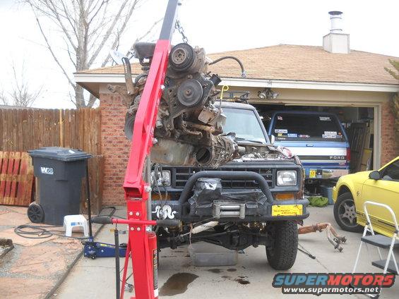 1992 Ford Explorer with engine removed, engine hoist lifting engine out. Blue Ford truck and yellow Mustang in background