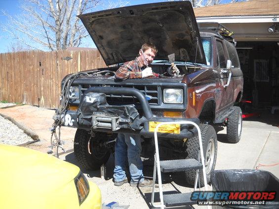 Man working on lifted brown Ford Bronco II with hood open, yellow car in foreground