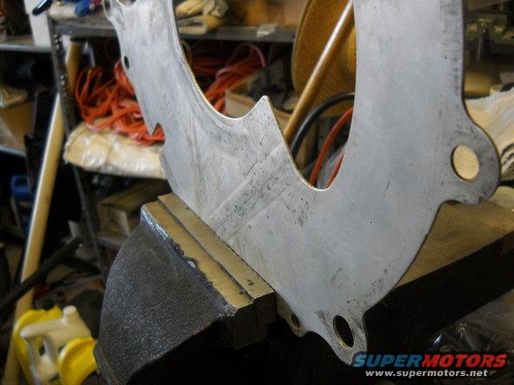 Metal bracket clamped in a vise, with "SUPERMOTORS.NET" visible at the bottom right