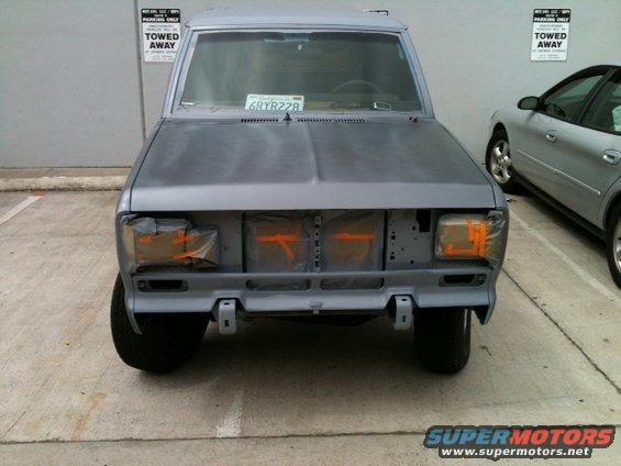 Front view of an SUV, unpainted, with orange tape on headlights and grille area. Another car is parked to the right