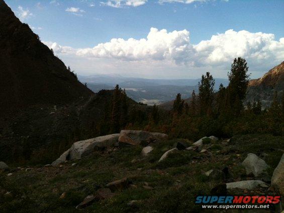 Mountain landscape with trees, rocks, and clouds, with a "SUPERMOTORS" logo in the lower right corner