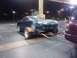 2003 Hyundai Tiburon - A dark blue coupe is loaded onto a trailer, hitched to a maroon SUV, at a gas station after dark.