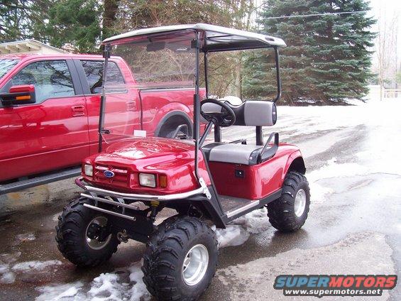 Red golf cart with oversized tires parked next to a red pickup truck. Snow on the ground