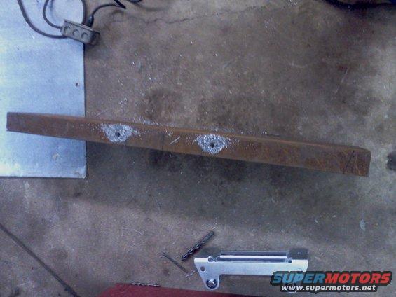 Metal bracket with two holes drilled into it, lying on a concrete floor next to a drill bit