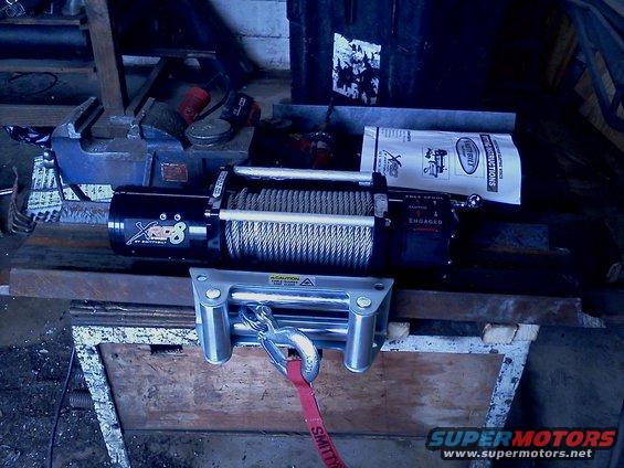 Winch with steel cable and a red strap attached to a hook, resting on metal bars over a wooden surface. A vise is visible to the left